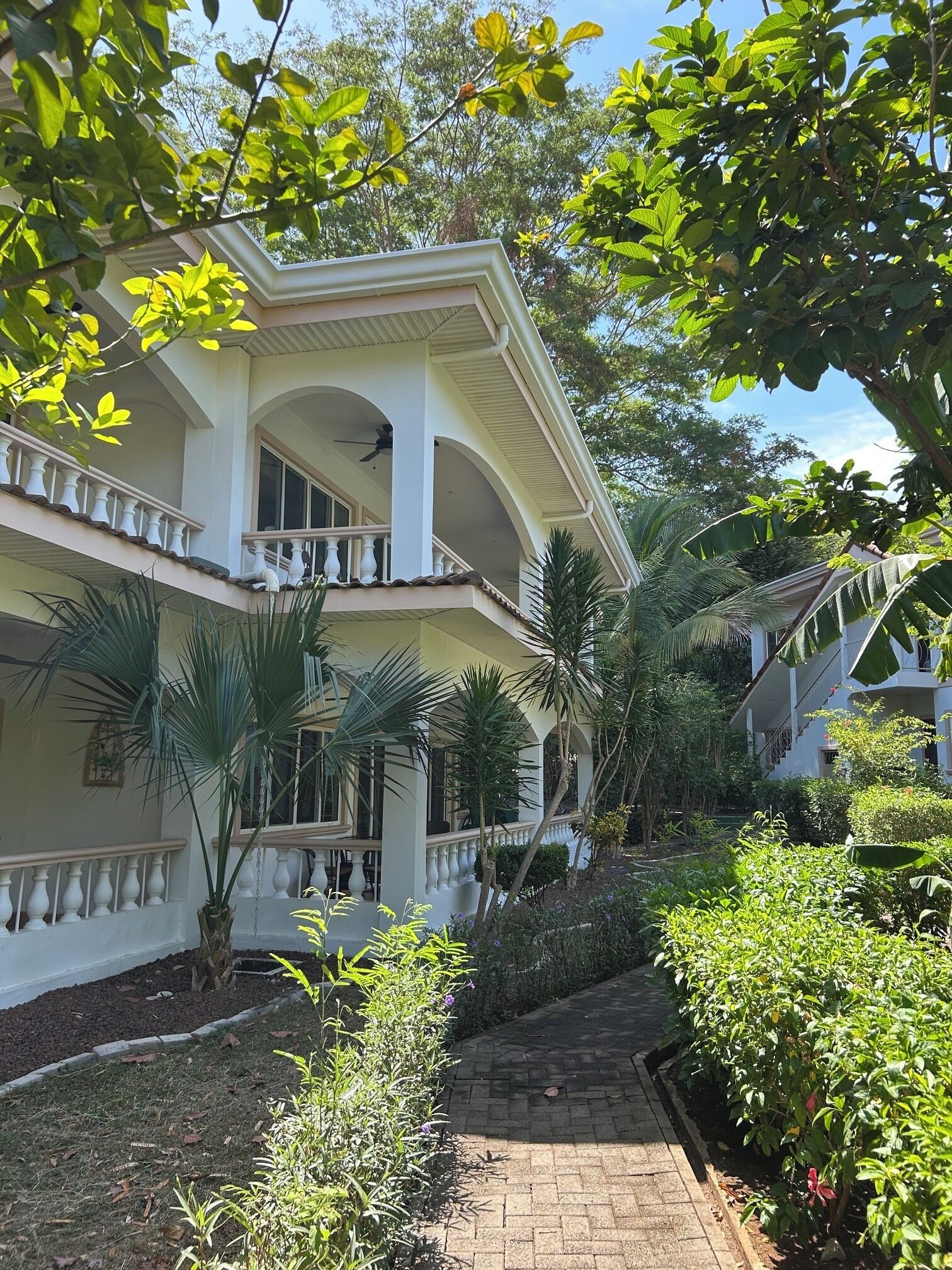 A two-story building with a balcony, surrounded by lush greenery and a paved pathway.