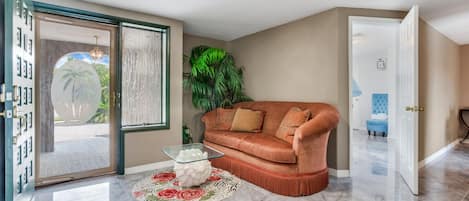 A living room with a sofa, a glass coffee table, a patterned rug, and a door leading to another room.