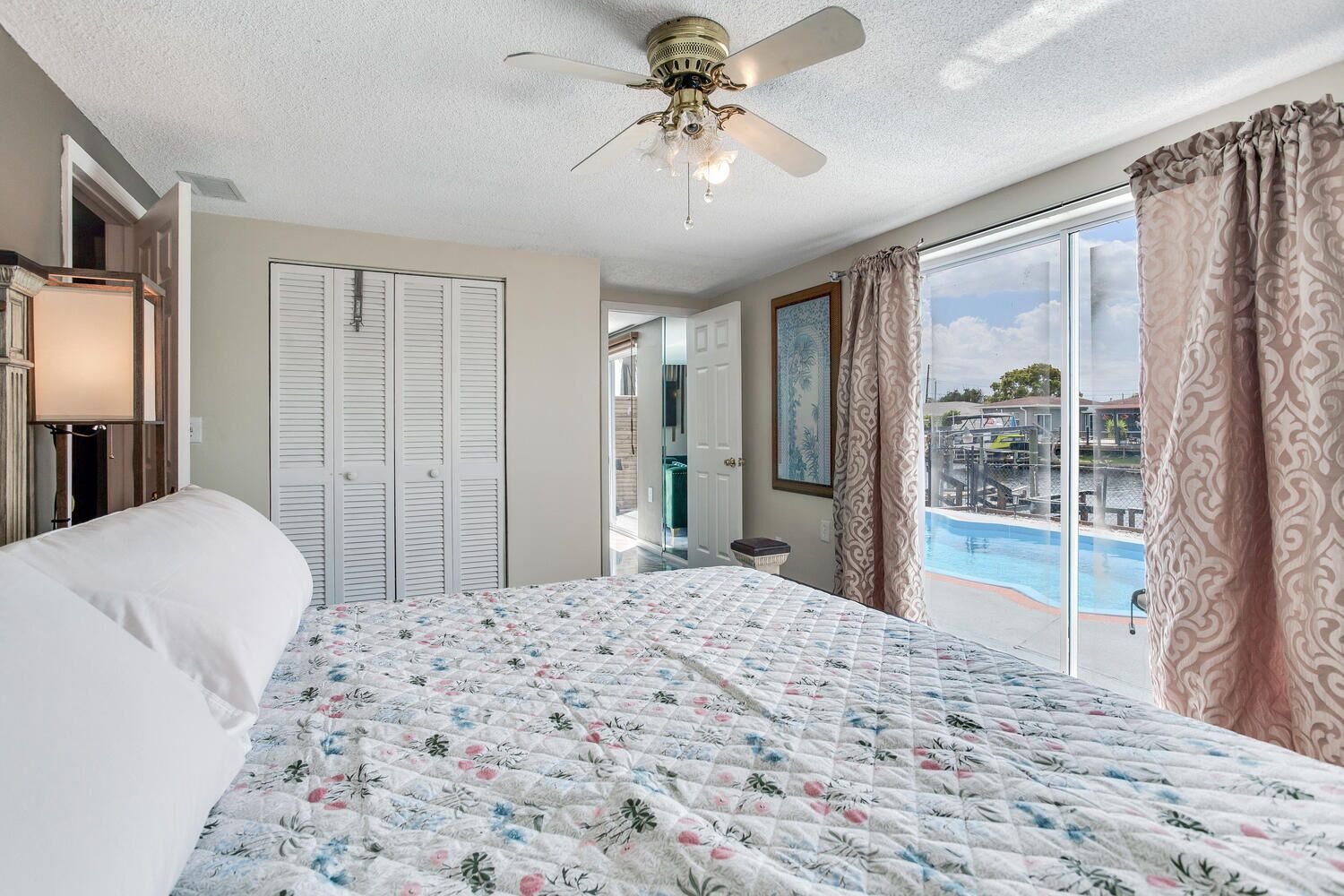 A bedroom with a bed, a ceiling fan, a view of a pool, and a door leading to a bathroom.