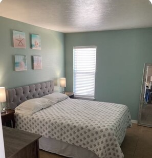 A bedroom with a bed, two bedside tables, a window with blinds, and wall art. - First Floor 2- Bedroom Bayside Condo (Fort Walton Beach)