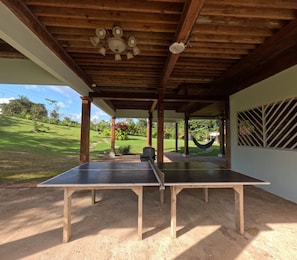 An outdoor ping pong table under a wooden pergola with a view of a grassy landscape and a hammock.