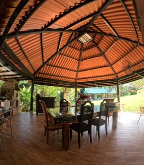 An outdoor covered dining area with a wooden table and chairs, a grill, and a view of a house in the background. - Finca Los Caobos (El Carrizal)