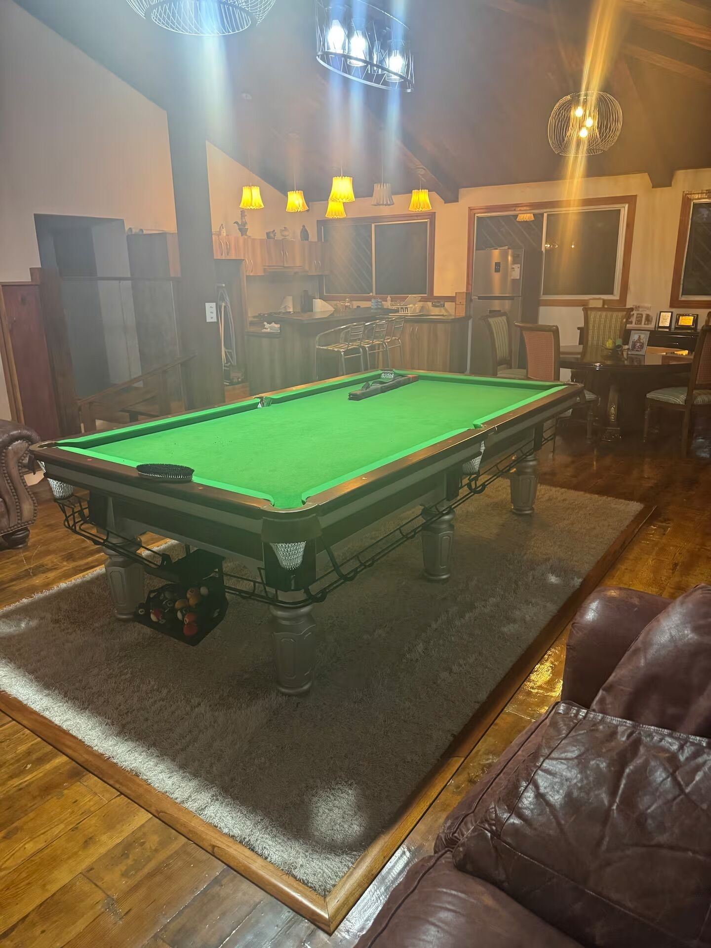 A pool table in a room with a wooden floor, a green felt surface, and a leather couch in the foreground.