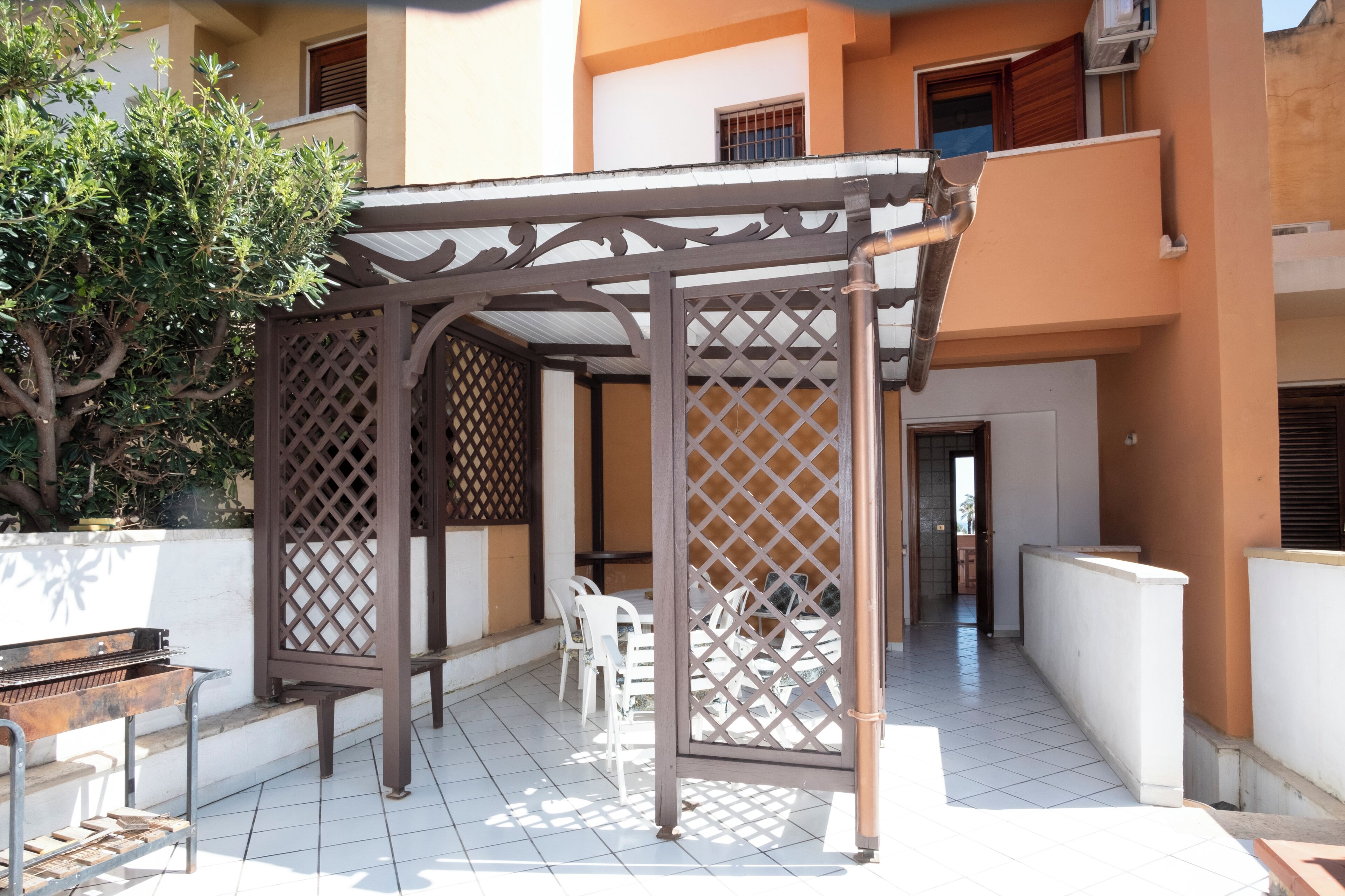 A covered outdoor patio with a grill, white chairs, and a wooden pergola.