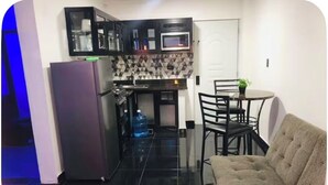 Cocina compacta con refrigerador, microondas y salpicadero de azulejos. Hay una zona de comedor con mesa y sillas, y un sofá.