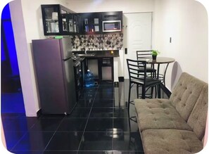 Eine kompakte Küche mit Kühlschrank, Mikrowelle und einer gefliesten Spritzschutzwand. Es gibt eine Essecke mit Tisch und Stühlen sowie eine Sitzecke mit einer Couch.