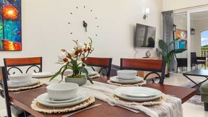Een eetruimte met een houten tafel gedekt voor vier personen, omringd door stoelen, een klok aan de muur en een televisie boven de tafel bevestigd.