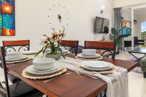 A dining area with a wooden table set for four, surrounded by chairs, a clock on the wall, and a television mounted above the table. - Richmond Estate 3rd floor condo~ Pool~Beach Access (Claremont)