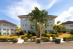 Un edificio residencial de varios pisos con fachada simétrica, rodeado de jardines cuidados y palmeras.