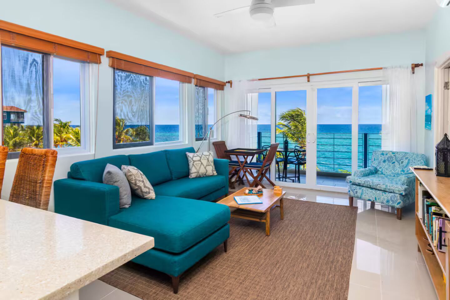 A room with a teal sofa, a dining table, and a view of the ocean through large windows.