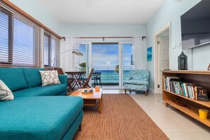 A living room with a sofa, a coffee table, a chair, a bookshelf, and a view of the ocean. - Teal Horizon~OceanfrontCaribbeanEscape - Tower Isle (Tower Isle)