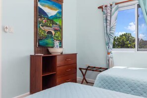 A bedroom with a bed, a wooden dresser with a bowl on top, a window with curtains, and a wall with a painting. - Teal Horizon~OceanfrontCaribbeanEscape - Tower Isle (Tower Isle)