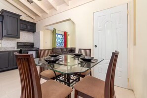 A dining area with a glass-top table surrounded by chairs, a kitchen in the background, and a door to the right. - 8BedRm Oasis ~Draxhall ~Pool ~Gym ~PlyGrnd ~Tennis (Saint Ann's Bay)