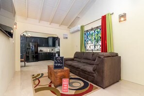 A living room with a sofa, a coffee table, and a rug. There is a kitchen in the background with dark cabinets and a window with curtains. - 8BedRm Oasis ~Draxhall ~Pool ~Gym ~PlyGrnd ~Tennis (Saint Ann's Bay)