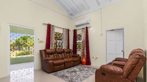 A living room with a brown sofa and a brown armchair, a patterned rug, a window with red curtains, and an air conditioning unit. - 3BR Oasis in Drax hall ~ Pool~ Gym ~ Gated ~Tennis (Saint Ann's Bay)