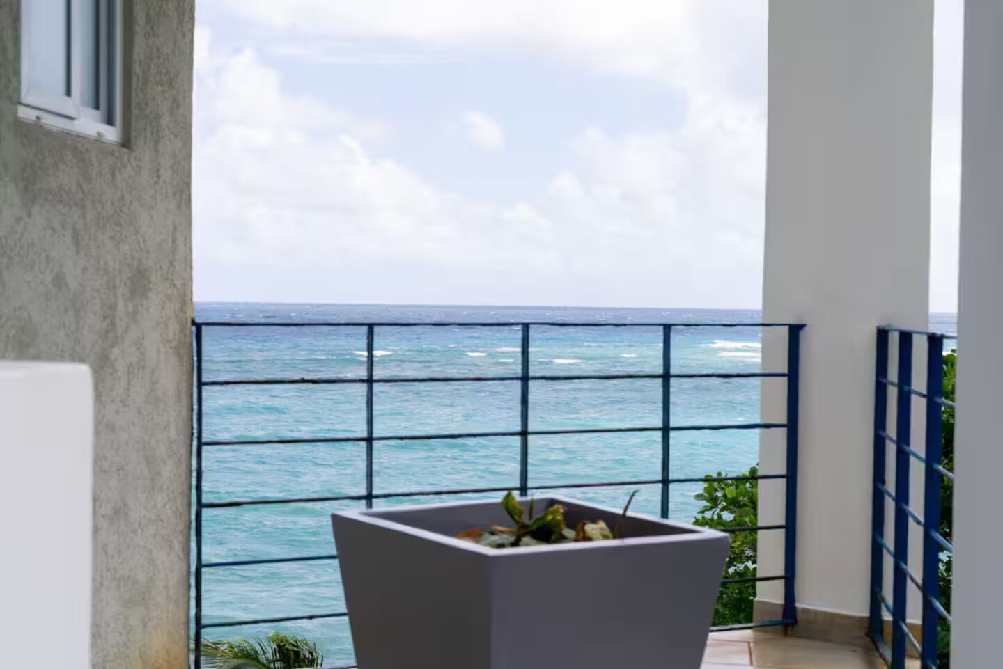 A balcony with a view of the ocean, a white planter with plants, and a blue railing.