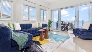 Ein modernes Wohnzimmer mit blauer Sofagarnitur, einem Couchtisch und Meerblick durch große Fenster.