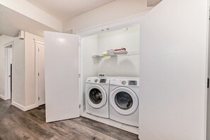 A laundry room with two front-loading washing machines, a shelf with folded clothes, and a door leading to another room. - Luxurious Basement: Hidden Oasis (Edmonton)