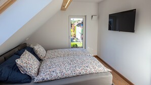 Ein Schlafzimmer mit schrägen Wänden, einem Bett mit floralem Bettzeug, einem Fernseher an der Wand und Blick auf einen Garten durch eine Tür.