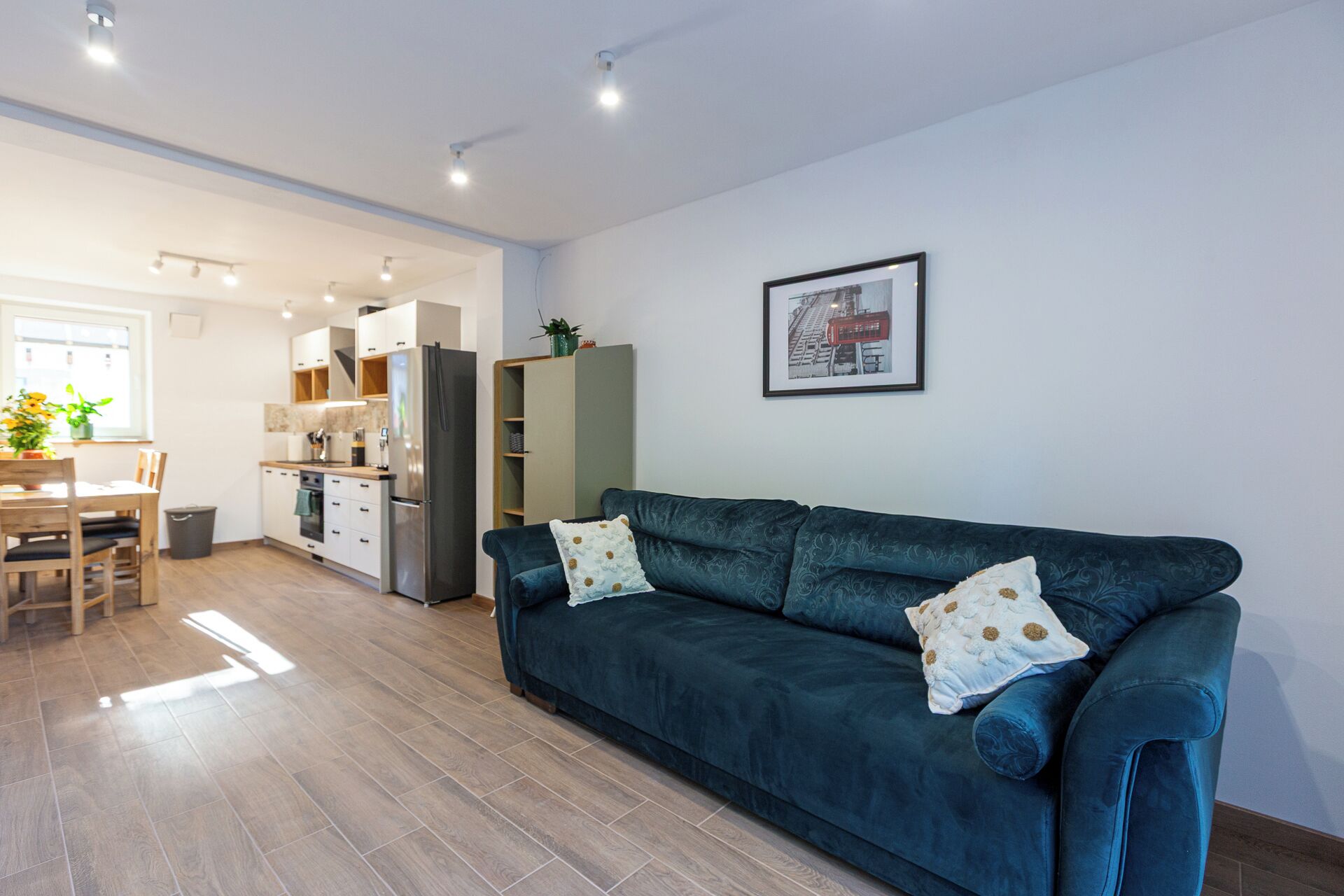 Ein modernes Wohnzimmer mit einem dunkelblauen Sofa, einer Essecke mit Holztisch und Stühlen, einer Küche mit weißen Schränken und einem Kühlschrank sowie einem gerahmten Bild an der Wand.
