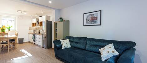Ein modernes Wohnzimmer mit einem dunkelblauen Sofa, einer Essecke mit Holztisch und Stühlen, einer Küche mit weißen Schränken und einem Kühlschrank sowie einem gerahmten Bild an der Wand.