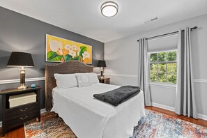 Ein Schlafzimmer mit einem Bett, Nachttischen, einem Gemälde, einem Fenster mit Vorhängen und einem gemusterten Teppich.