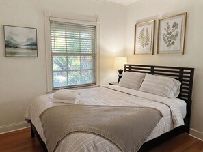 Ein Schlafzimmer mit Bett, einem Fenster mit Jalousien, einer Nachttischlampe und gerahmten Pflanzenbildern an der Wand.