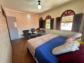 Ein Schlafzimmer mit einem Bett, einem Sessel, einem Ventilator und einem Fenster mit Rollläden.