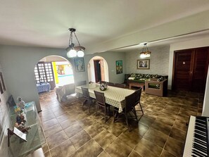 A dining area with a glass table, chairs, and a piano. There are arched doorways and a sofa in the background. - Beach house with pool in Porto das Dunas, 7 bedrooms + garage for 6 cars. (Aquiraz)