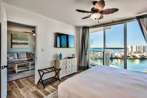 A modern hotel room with a large bed, a ceiling fan, a TV, and a view of the city and water. - Destin’d to Unwind | Walk to Beach | Gulf Views (Destin)