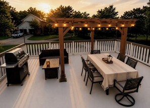 An outdoor patio with a pergola, string lights, a grill, a table with chairs, and a view of a house in the background.