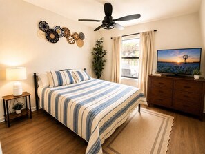 A bedroom with a bed, a television, a ceiling fan, a window with curtains, and wall decorations.