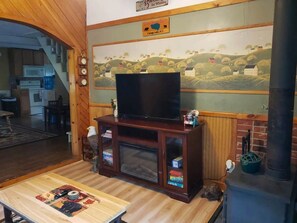A living room with a television on a wooden cabinet, a fireplace, and a mural on the wall. - Cozy Adirondack Woodland Retreat- near Caroga Lake (Caroga Lake)