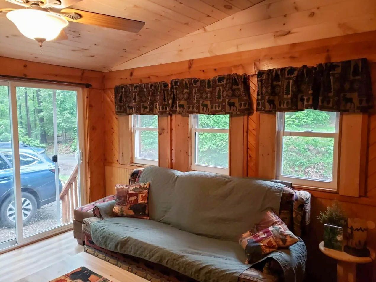 A cozy cabin interior with a sofa, patterned curtains, and a view of the outdoors through large windows.