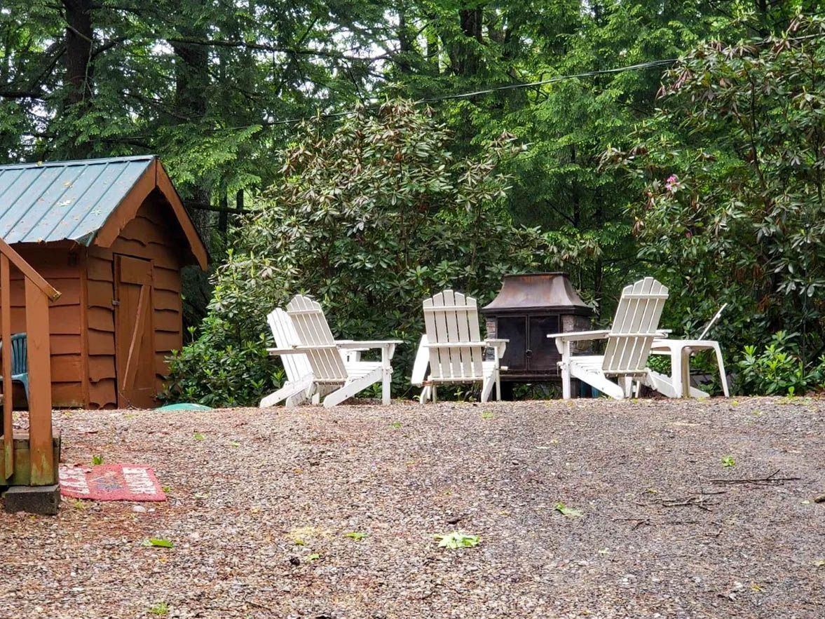 A wooden cabin with a metal roof, surrounded by dense foliage. There are four white chairs arranged in a semi-circle facing a small fireplace.