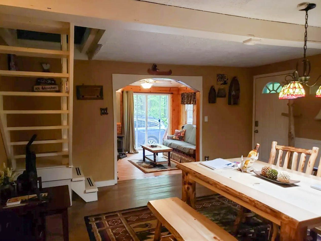 Ein Essbereich mit einem Holztisch und Bänken, einem Bücherregal und durch einen Bogen hindurch einem Blick in ein Wohnzimmer mit einer Couch und einem Fenster.