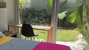 Une chambre à coucher avec un lit, un bureau et une chaise. Une grande fenêtre offre une vue sur un jardin.