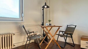 Een kamer met een radiator, een raam, een tafel met twee stoelen, een lamp en een kleine plant.