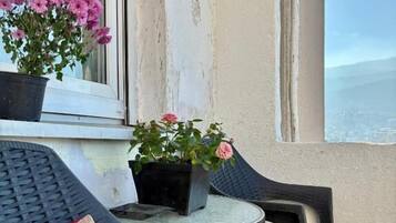 En balkon med et rundt bord og to stole, en potteplante med lyserøde blomster, og havudsigt.