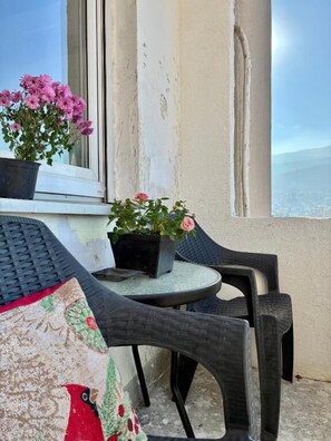 Un balcón con una mesa redonda y dos sillas, una planta en maceta con flores rosas, y vista al mar.