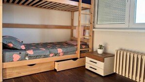 Een stapelbed met een onderbed, een nachtkastje met lades, een radiator en een raam met zonwering.
