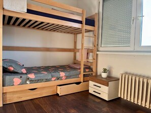 Una cama litera con una cama inferior, una mesita de noche con cajones, un radiador y una ventana con persianas.