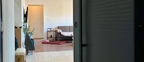 Un couloir mène à une pièce dont la porte est fermée. On y aperçoit un tapis à motifs et une chaise au fond de la pièce.
