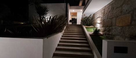 Une vue nocturne d’un escalier moderne menant à l’entrée d’un bâtiment, entouré de plantes et de murs en pierre.
