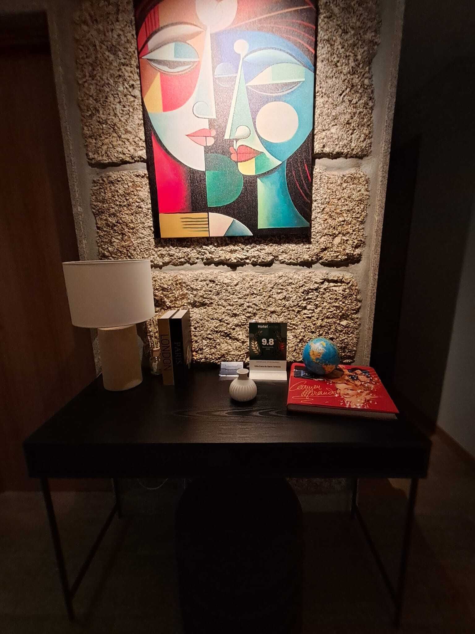 Habitación de hotel con pared de piedra, un cuadro, una lámpara, un globo terráqueo y un libro rojo sobre una mesa.