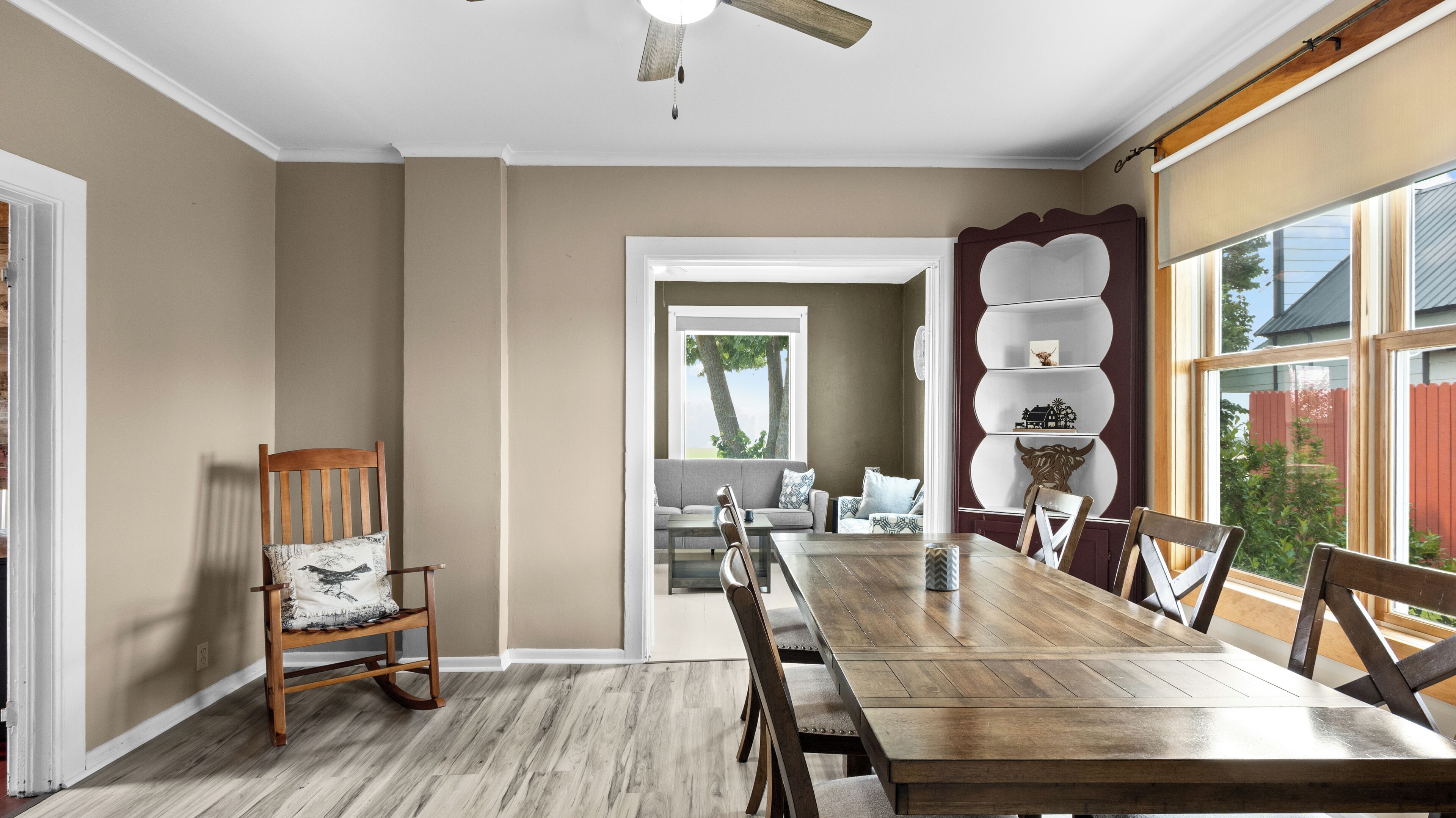 Une salle à manger avec une table en bois, des chaises, un fauteuil à bascule et, vue à travers une porte, un salon.