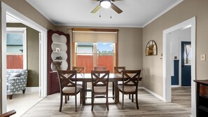 Une salle à manger avec une table et des chaises en bois, un ventilateur de plafond et une vue sur une cour clôturée.