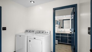 Un lavadero con dos lavadoras y secadoras frontales blancas, una puerta azul que da a un baño con lavamanos y espejo.