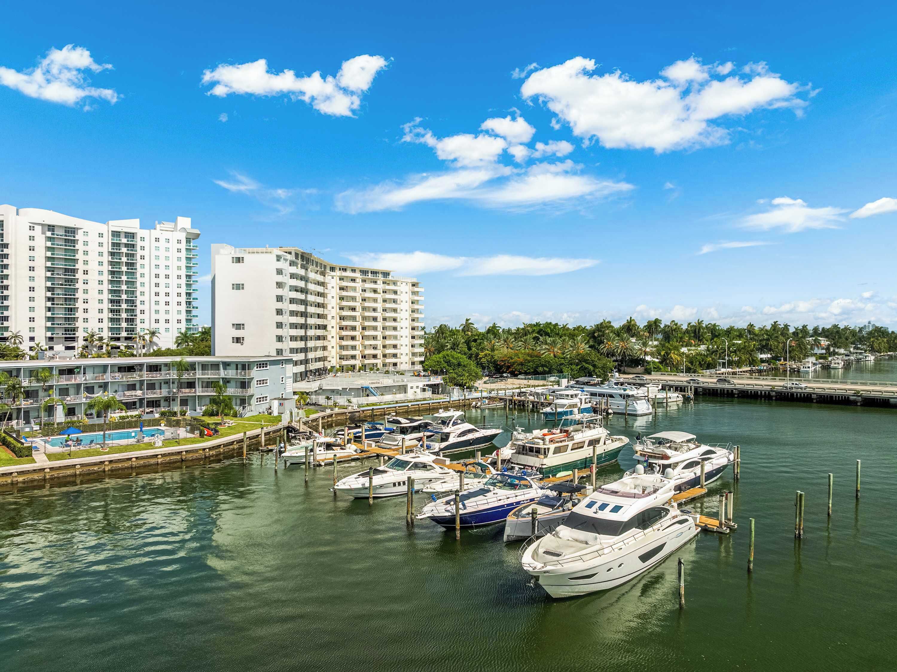 A marina with multiple boats docked, a large hotel building, and a clear blue sky.