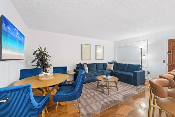 A modern living room with a blue sofa set, a wooden coffee table, and a dining table with chairs. - Bahia Serena - Bay Views on Marina With Parking (North Bay Village)
